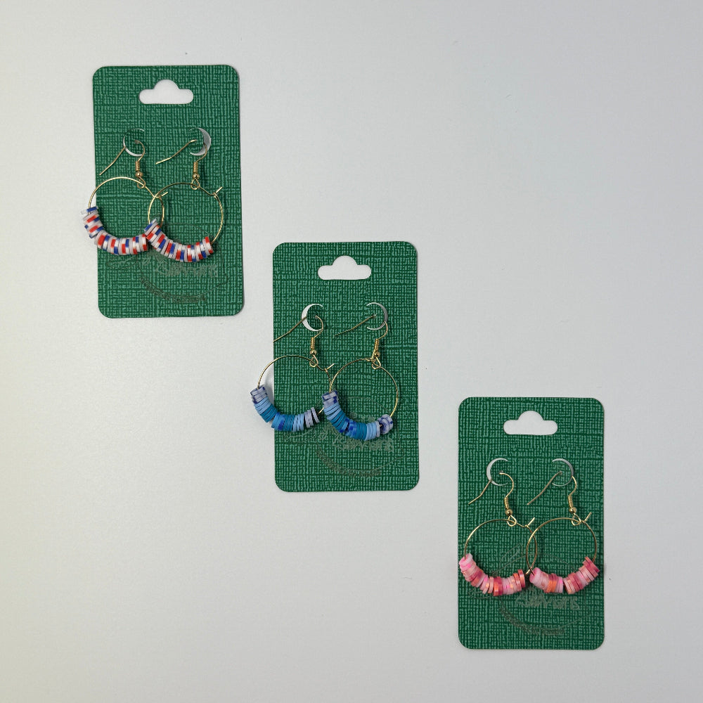 Three pairs of clay beed hoop earrings on green cards against a light gray background